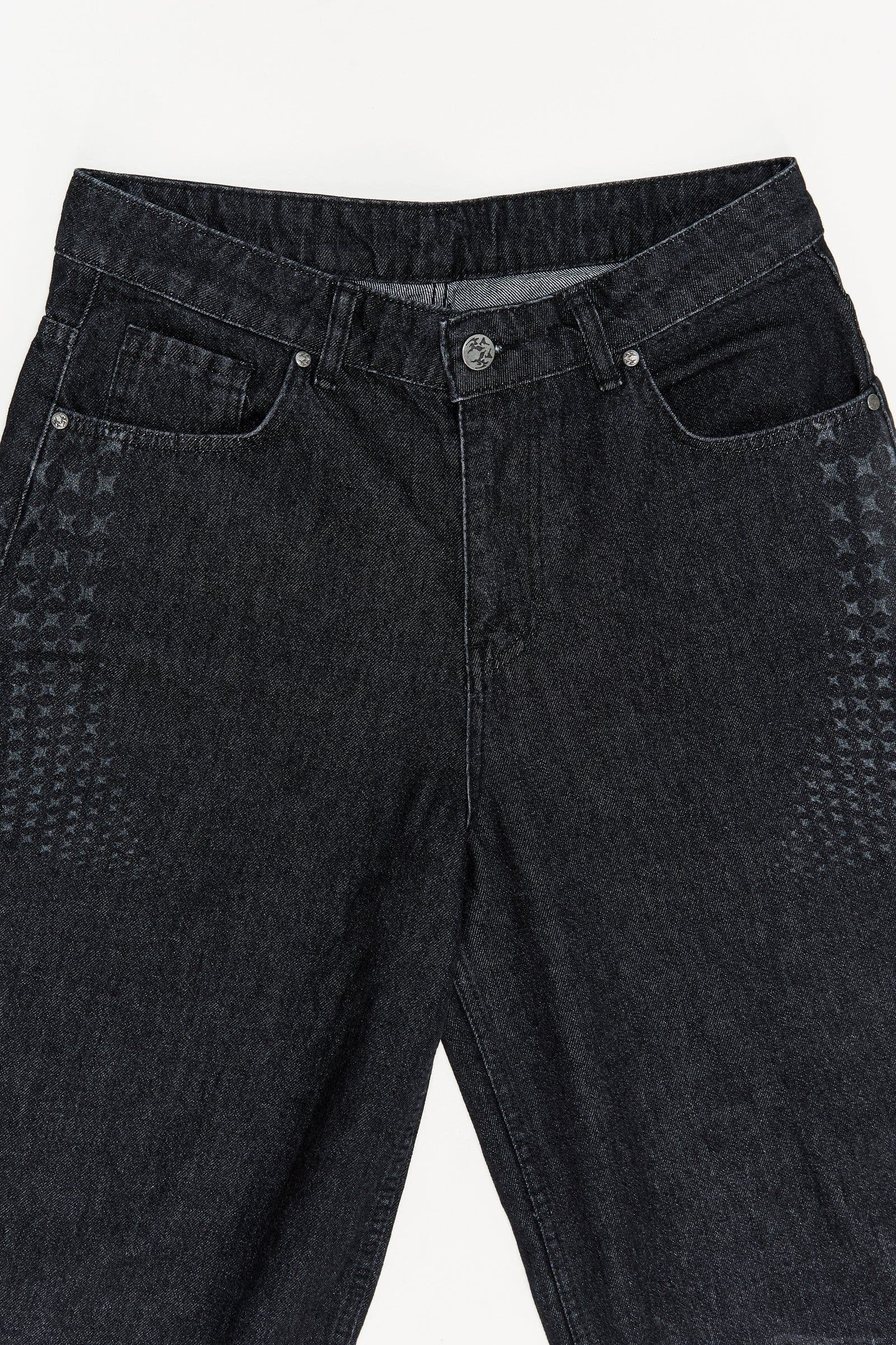 Close view of dark jeans laid flat, showing the waistband, front pockets and small repeated motifs along the sides
