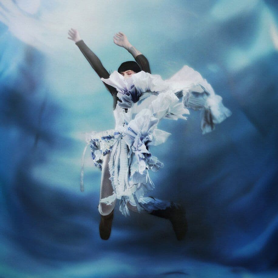 Figure in torn white fabric with raised arms against a blurred blue background